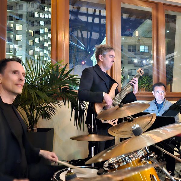 A jazz band performs indoors with drums, bass guitar, and keyboard.