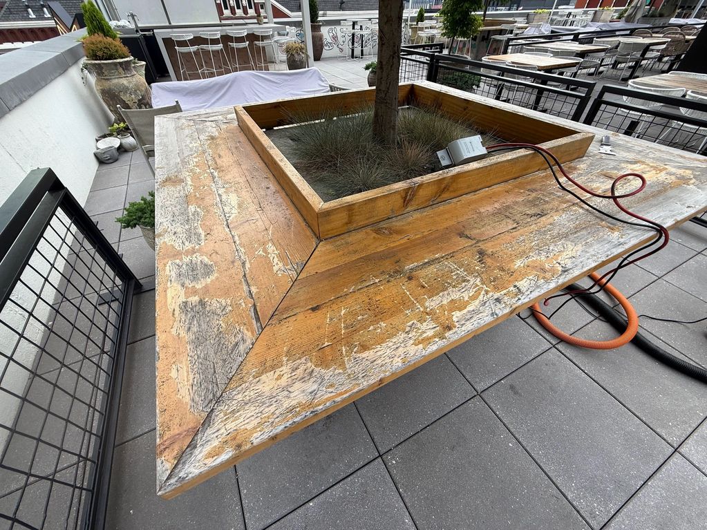 Weathered wooden table with a tree planter in the center on a tiled patio.