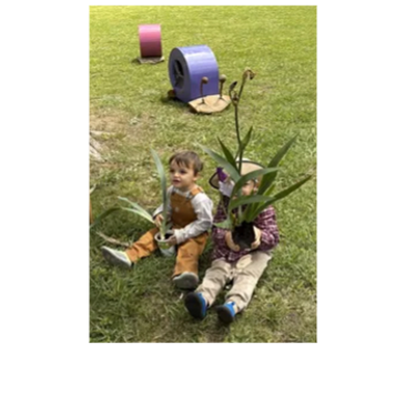Two toddlers sitting on grass holding potted plants with colorful garden decorations behind them.