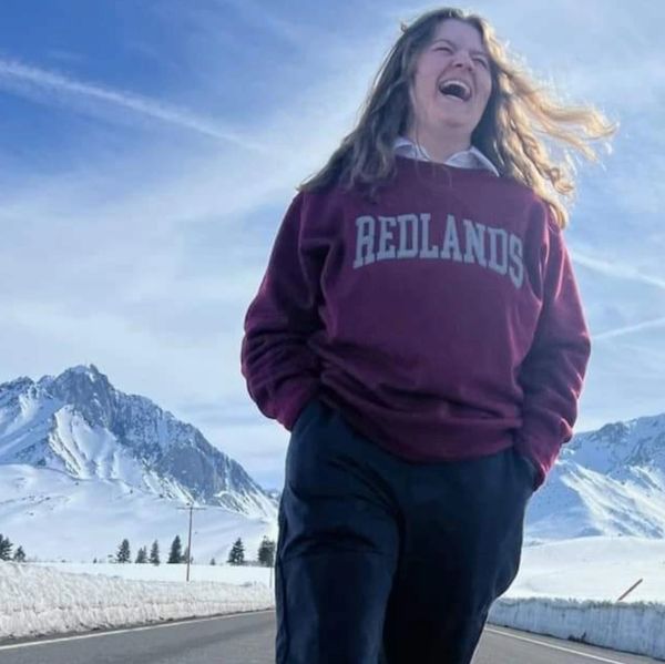 A joyful woman laughs outdoors with snowy mountains behind her.
