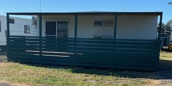 Small prefabricated building with a green porch fence on grassy ground.