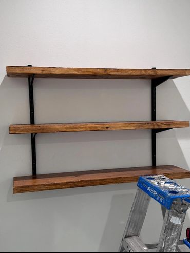 Three wooden shelves mounted on a wall with a ladder in front.