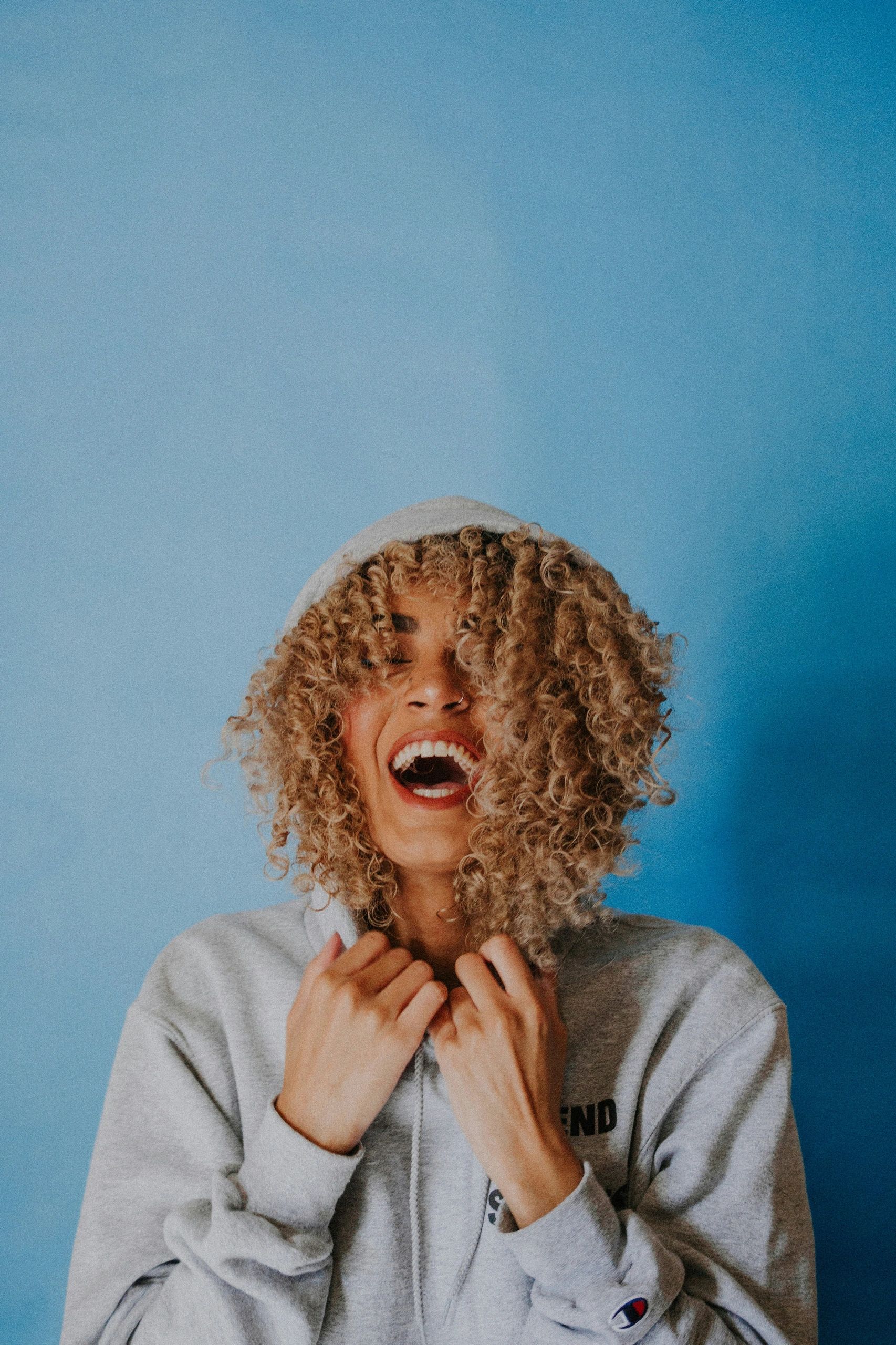 Joyful woman with curly hair laughing in a gray hoodie against a blue background.