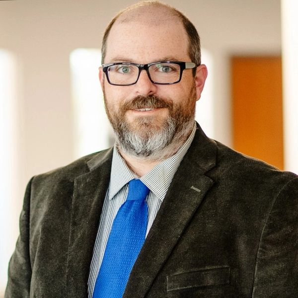 Smiling man in glasses and blue tie wearing a dark blazer.