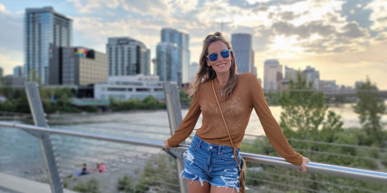 Woman in sunglasses and casual outfit posing on a bridge with city skyline in the background.