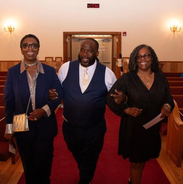 Three people smiling and walking arm in arm down a church aisle.