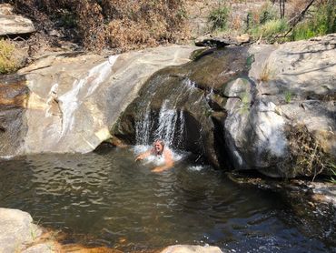 A smiling man swims in a creek pool with water cascading onto his head from the creek above.