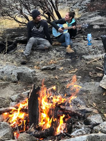A young man and woman play guitar and sing by a small campfire. 