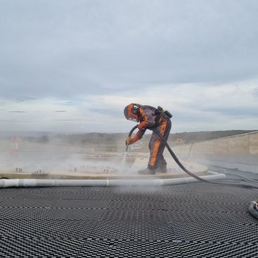 Worker in protective gear spraying a surface with high-pressure hose outdoors.
