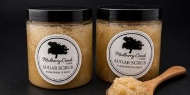 Two jars of Mulberry Creek Sugar Scrub with a wooden scoop of scrub.