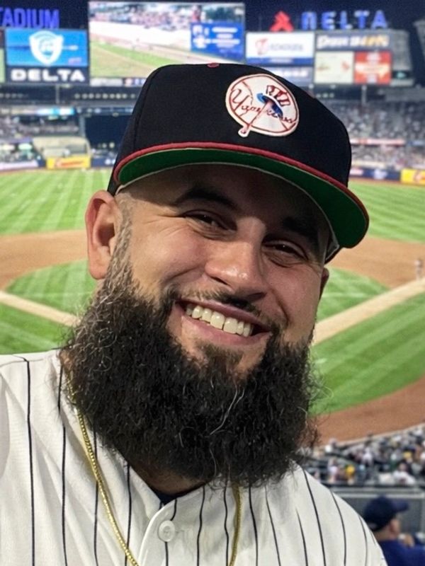 A happy man with a thick beard at a baseball stadium.