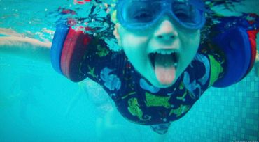 Child underwater in pool with tongue out and wearing goggles and floaties.