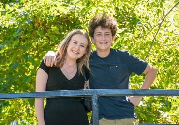 Two smiling teenagers posing outdoors with lush green trees in the background.