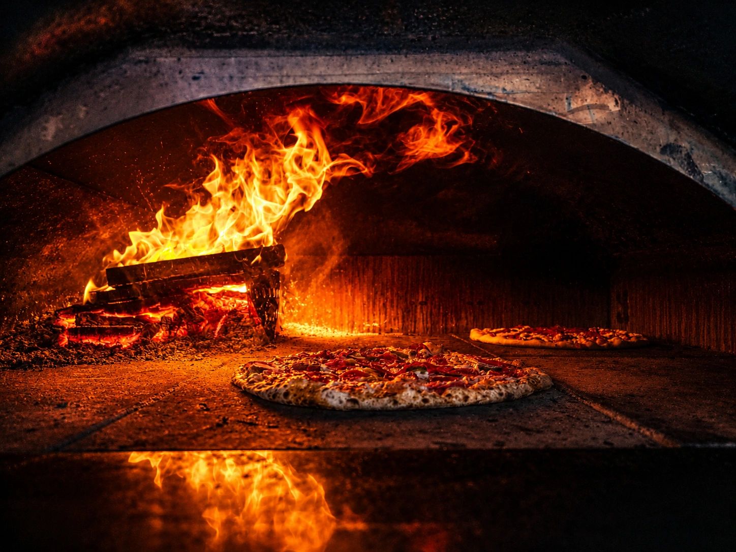 Two pizzas baking inside a wood-fired oven with flames.
