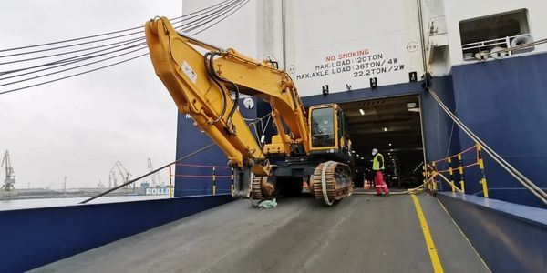 Excavator being loaded onto a ship via a ramp in a port area.