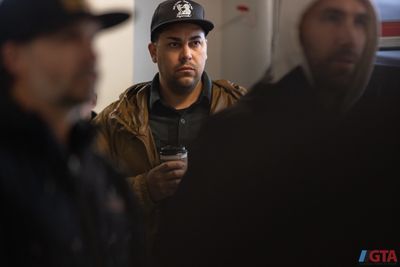 Man in a brown jacket and black cap holding a coffee cup indoors.