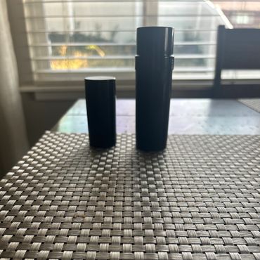 Two black cylindrical containers on a woven placemat near a window.