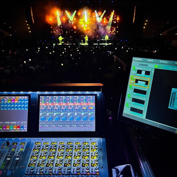 Sound mixing console and screens at a live concert with stage lights and performers.