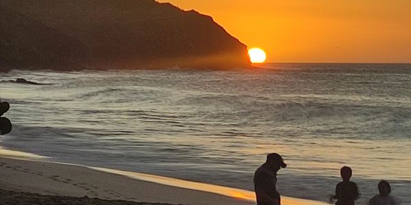 Silhouettes of people on a beach at sunset with the sun setting behind a mountain.