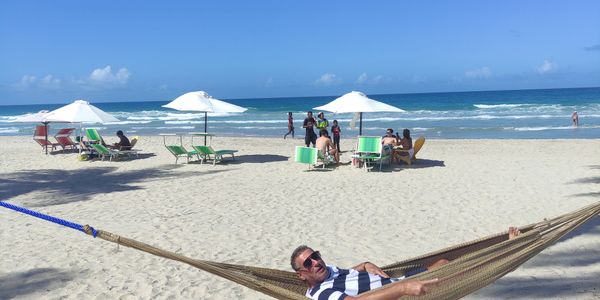 Man relaxing on a hammock at a sunny beach with umbrellas and people in the background.