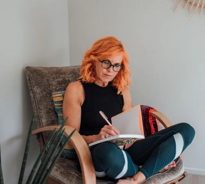 Woman with red hair writing in a notebook while seated in a cozy chair.