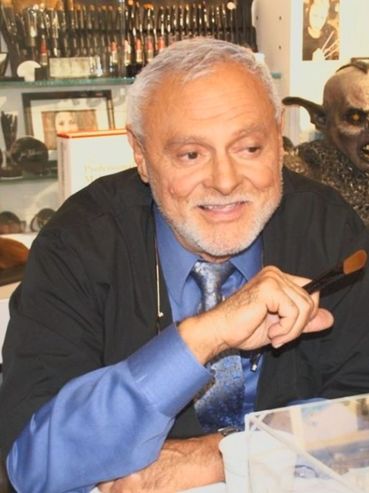 An older man smiling in a makeup studio with horror character masks behind him.