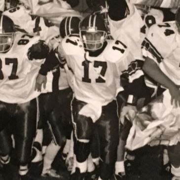 A high school football team runs onto the field through a banner in black and white.