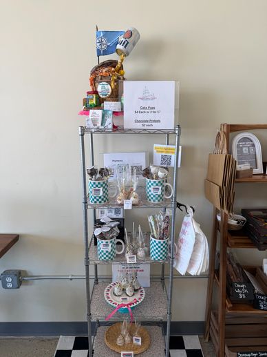 A metal shelf displays assorted mugs, treats, and signs for sale in a cozy shop corner.