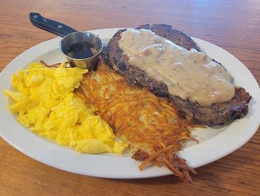 Steak with gravy, scrambled eggs, hash browns, and syrup on a white plate.