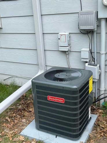 A Goodman air conditioning unit installed outside a house with gray siding.