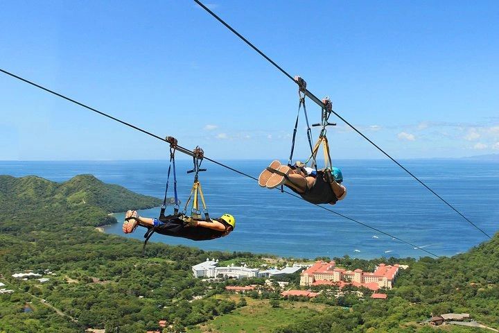 Dos personas en tirolesas sobre un paisaje con mar y montañas.
