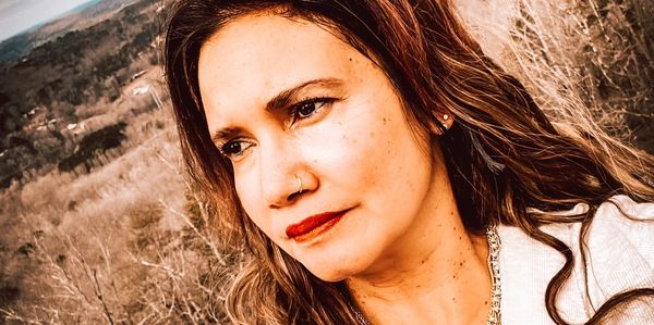 A woman with long hair and layered necklaces looks thoughtfully into the distance outdoors.