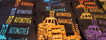 Stacks of black t-shirts with colorful DZ Automotive & Hauling LLC logos and truck illustrations.