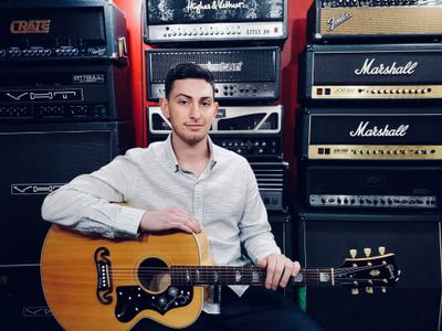 Man holding an acoustic guitar in front of stacked guitar amplifiers.