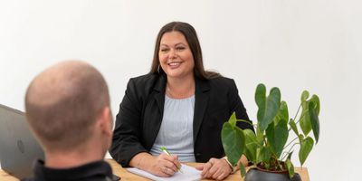 A woman smiles while talking to a man across a desk with a plant.