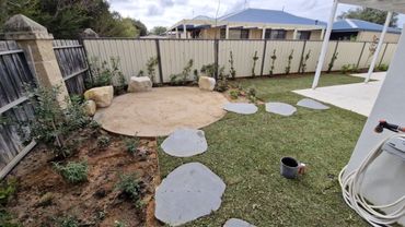 A backyard with stone path, small plants, and a circular sand area surrounded by rocks.