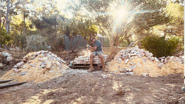 Man working with bricks and a power tool outdoors in the sun.