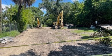 Construction site with excavators and caution tape in a sunny, tree-lined area.