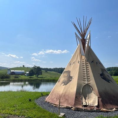 A tipi by a lake with a farm in the background.