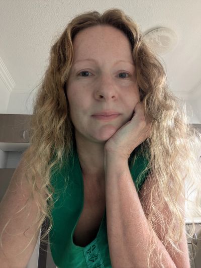 Woman with curly blonde hair resting her face on her hand, wearing a green top.
