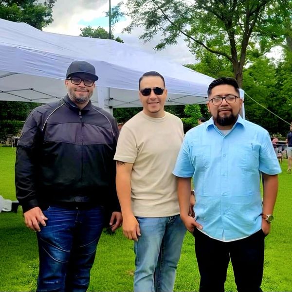 Three men posing outdoors at a park event under a white canopy.
