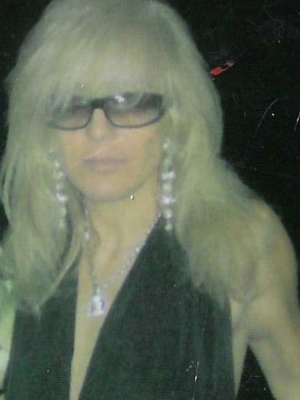 Woman with long blonde hair, glasses, and jewelry in a dark setting.