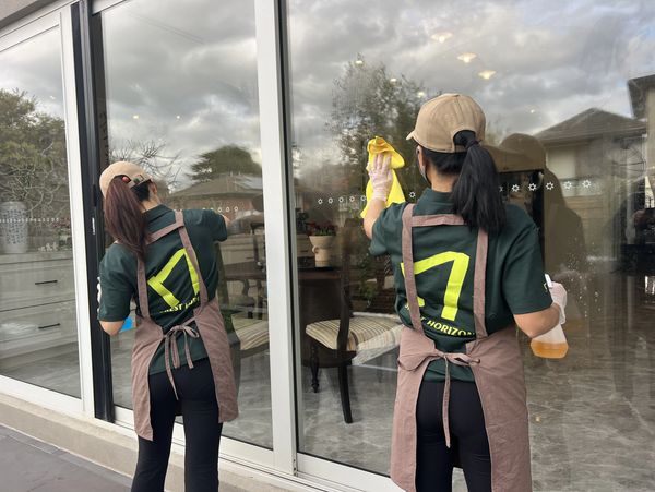 Two women cleaning large glass windows wearing aprons and caps.