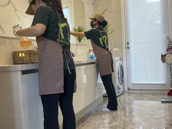 Two women cleaning a modern bathroom wearing aprons and caps.