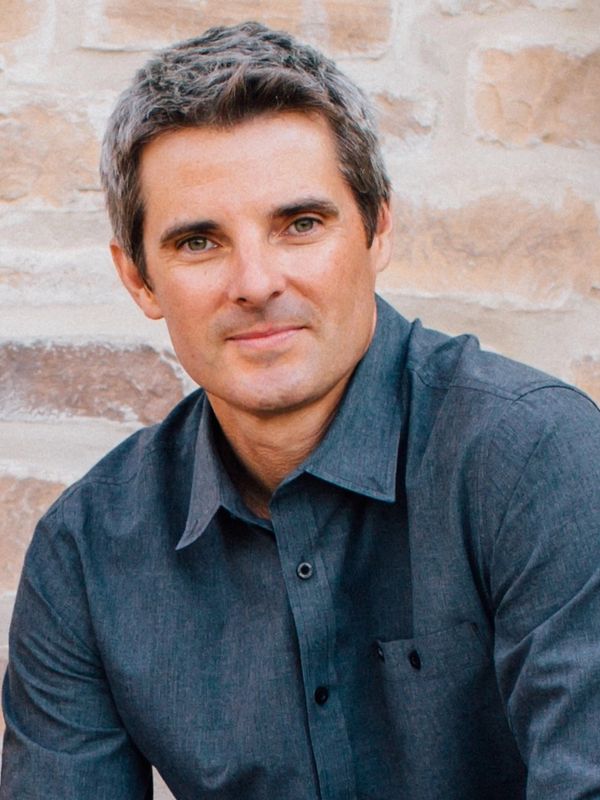 A man with gray hair wearing a dark blue shirt, posing against a stone wall.