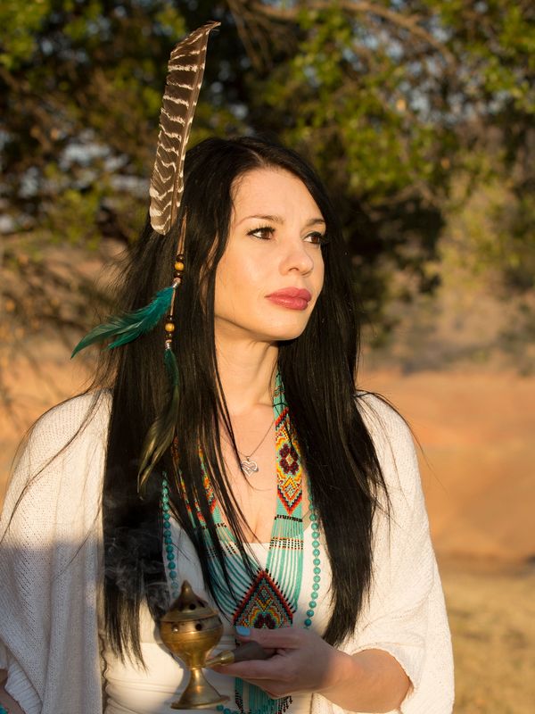 Woman with feathers in hair holding a smoking incense burner outdoors.