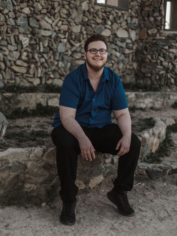 A smiling man in a blue shirt sits on a stone ledge outdoors.