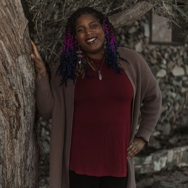 A smiling woman with colorful braided hair stands by a tree, wearing a maroon top and brown cardigan.