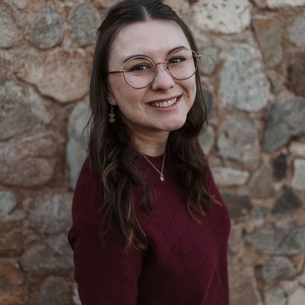 Smiling woman with glasses and maroon sweater against a stone wall.