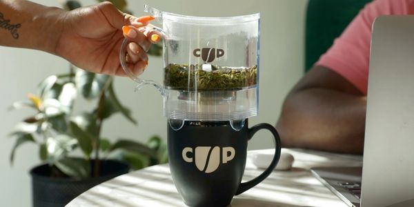 Hand holding tea infuser above black cup on table.